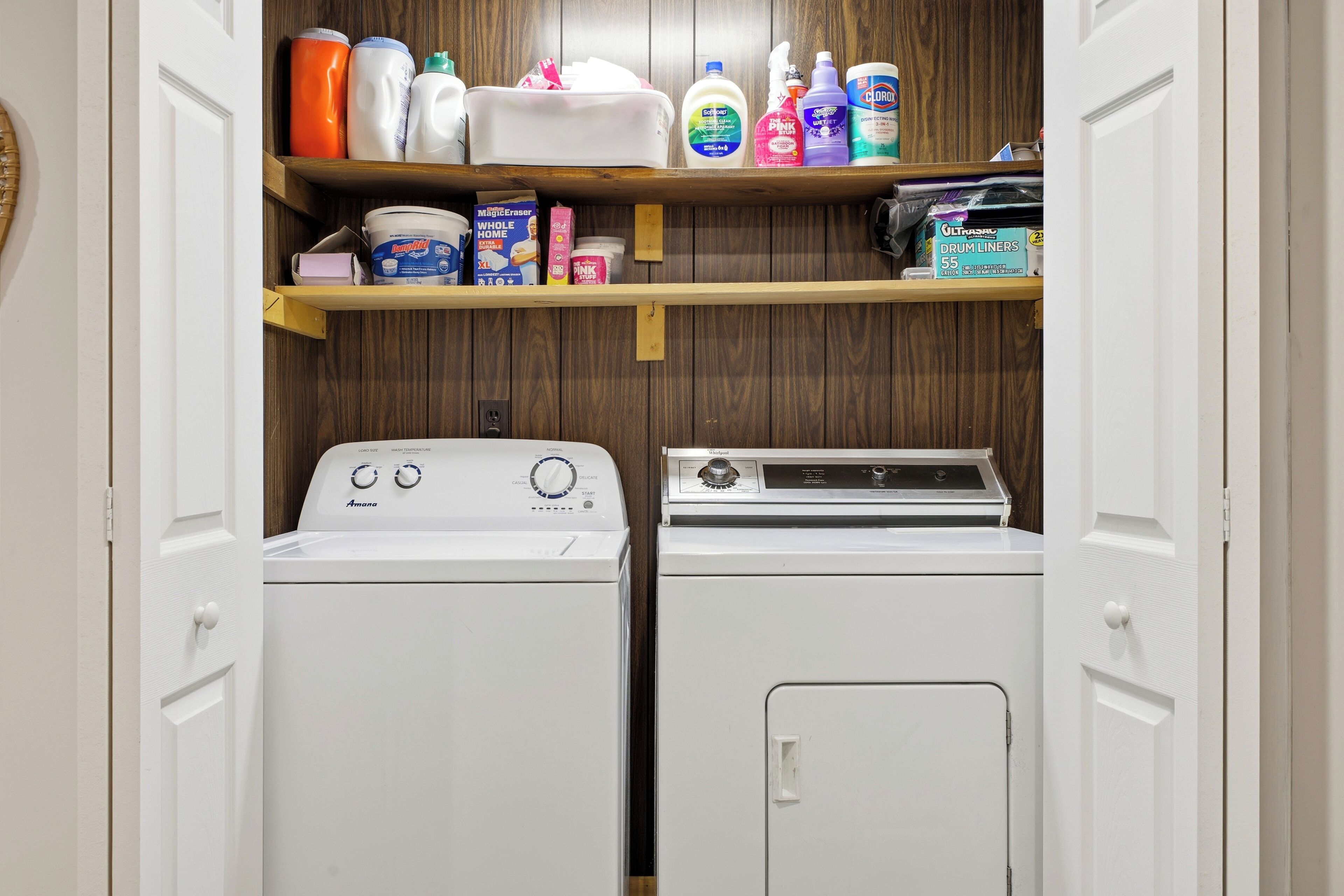 A laundry room with two white front-loading washers and dryers, wooden shelves above holding various cleaning products.