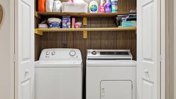 A laundry room with two white front-loading washers and dryers, wooden shelves above holding various cleaning products.