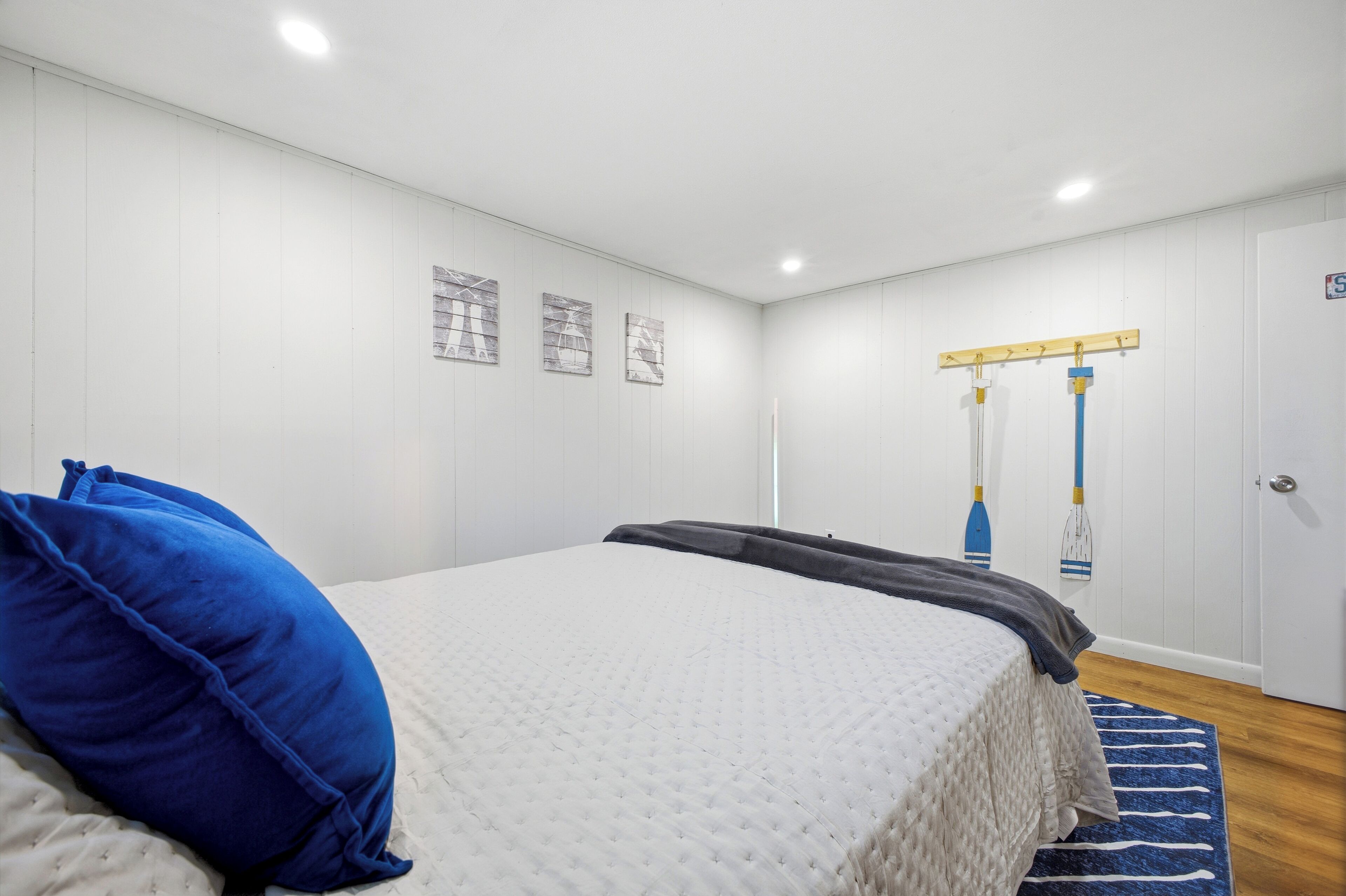 Un dormitorio con una cama, almohadas, un tapete azul y blanco a rayas, una pared blanca con tres cuadros enmarcados y una puerta con un cartel.