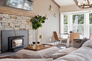 A cozy living room with a stone fireplace, a dining area with a table and chairs, and a large window with a view of trees.