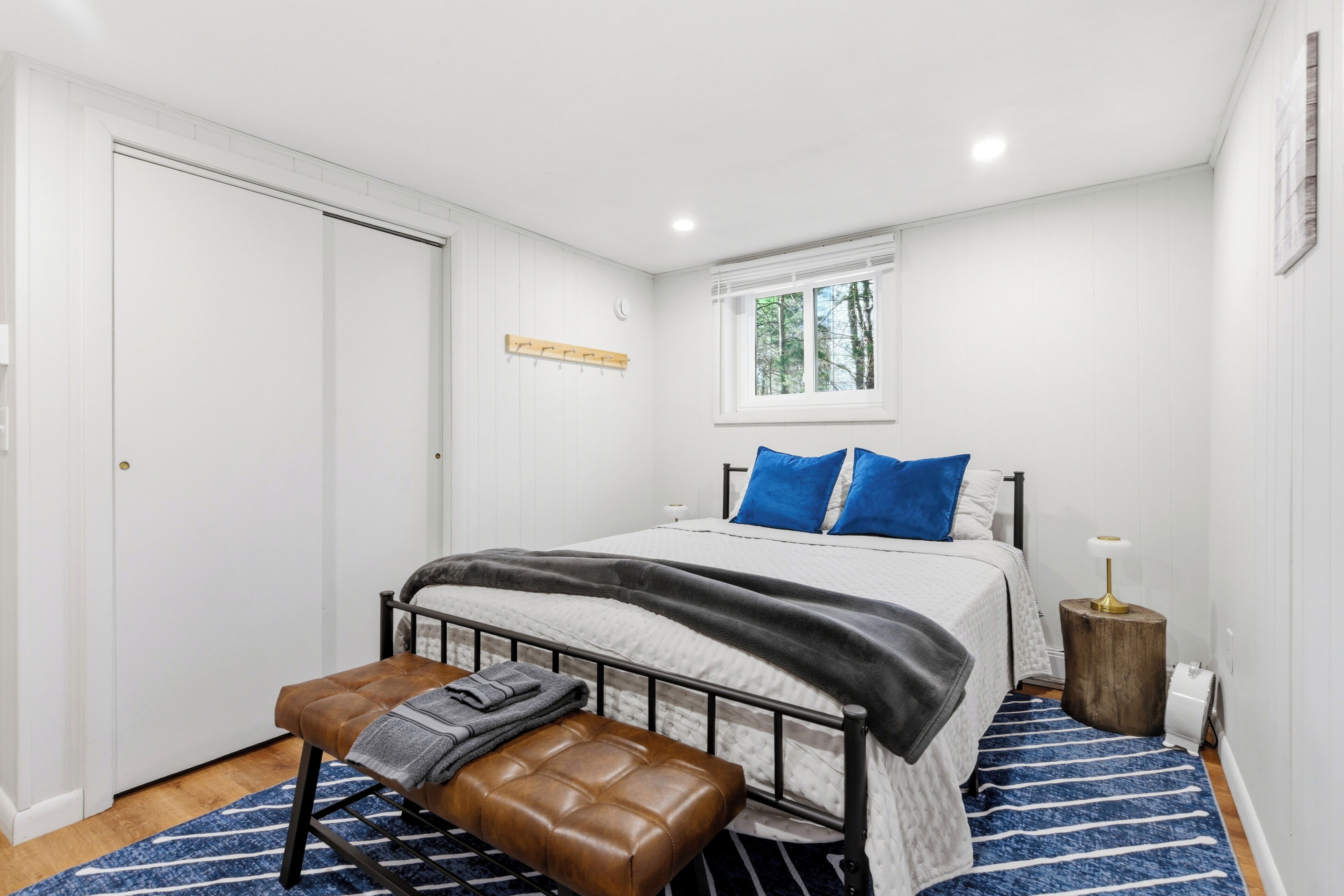 A bedroom with a bed, bench, bedside table, and a window.