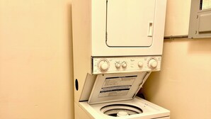 Lavadora blanca de carga frontal y compacta, con panel de control, ubicada en un rincón con piso de baldosas.