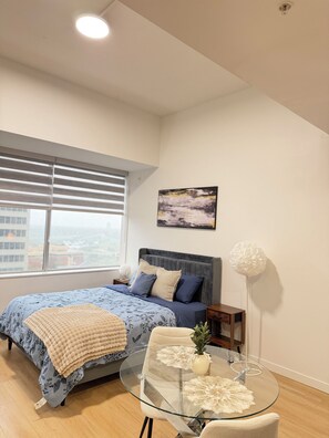 A bedroom with a bed, a glass table, a chair, a nightstand, a lamp, and a window with blinds. -     Urban Studio Oasis (Edmonton)