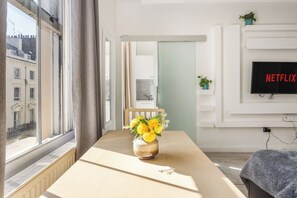 Ein Zimmer mit großem Fenster, einem Tisch mit Blumenvase, einem Sessel, einem Wand-Fernseher und einer Pflanze.
