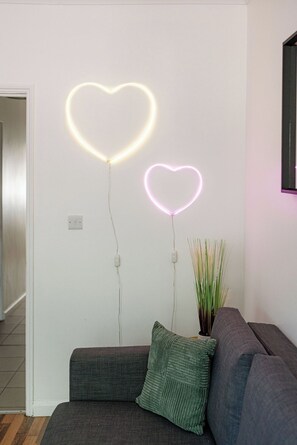 Ein modernes Wohnzimmer mit einem grauen Sofa, einem grünen Kissen und zwei herzförmigen Neonlichtern an der Wand.