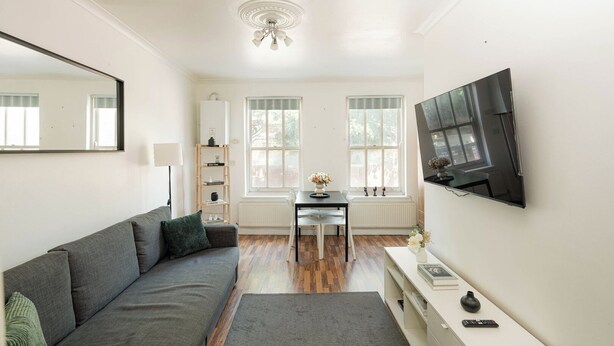 A modern living room with a grey sofa, a dining table, and a TV mounted on the wall. - Cozy Flat Near Station and Local Parks (London)
