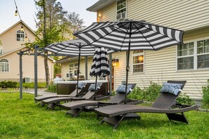 A patio with outdoor furniture, including lounge chairs and a striped umbrella, in front of a house with a hot tub. - Private Heated Pool | Hot Tub | Game Room (Blakeslee)