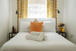Ein ordentlich bezogenes Bett mit weißer Bettwäsche, ein sternförmiges oranges Kissen und gerollte Handtücher. Es befinden sich zwei schwarze Nachttische mit Flaschen und eine Blumenvase darauf.