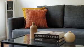 En moderne stue med en grå sofa, en gul og rød pude med struktur, et glas sofabord med en bog med titlen 'Leaders in Style at Home' og dekorative vaser.