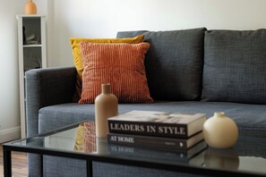 Ein modernes Wohnzimmer mit einem grauen Sofa, einem gelb-roten Kissen mit Struktur, einem Glastisch mit einem Buch mit dem Titel 'Leaders in Style at Home' und dekorativen Vasen.