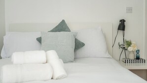 Ein ordentlich bezogenes Bett mit weißen Leinen und zwei Kissen, ein Nachttisch mit einer Lampe und einem Blumenstrauß, sowie eine Steckdose an der Wand.