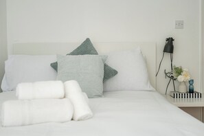 A neatly made bed with white linens and two pillows, a bedside table with a lamp and a vase of flowers, and a power outlet on the wall. - Bright Flat Near Parks and Restaurants (London)
