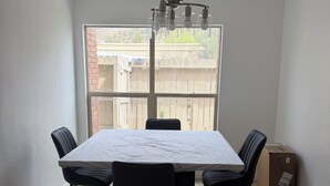 Un coin repas comprenant une grande table, quatre chaises, un luminaire au plafond et une fenêtre donnant sur une clôture et des arbres.