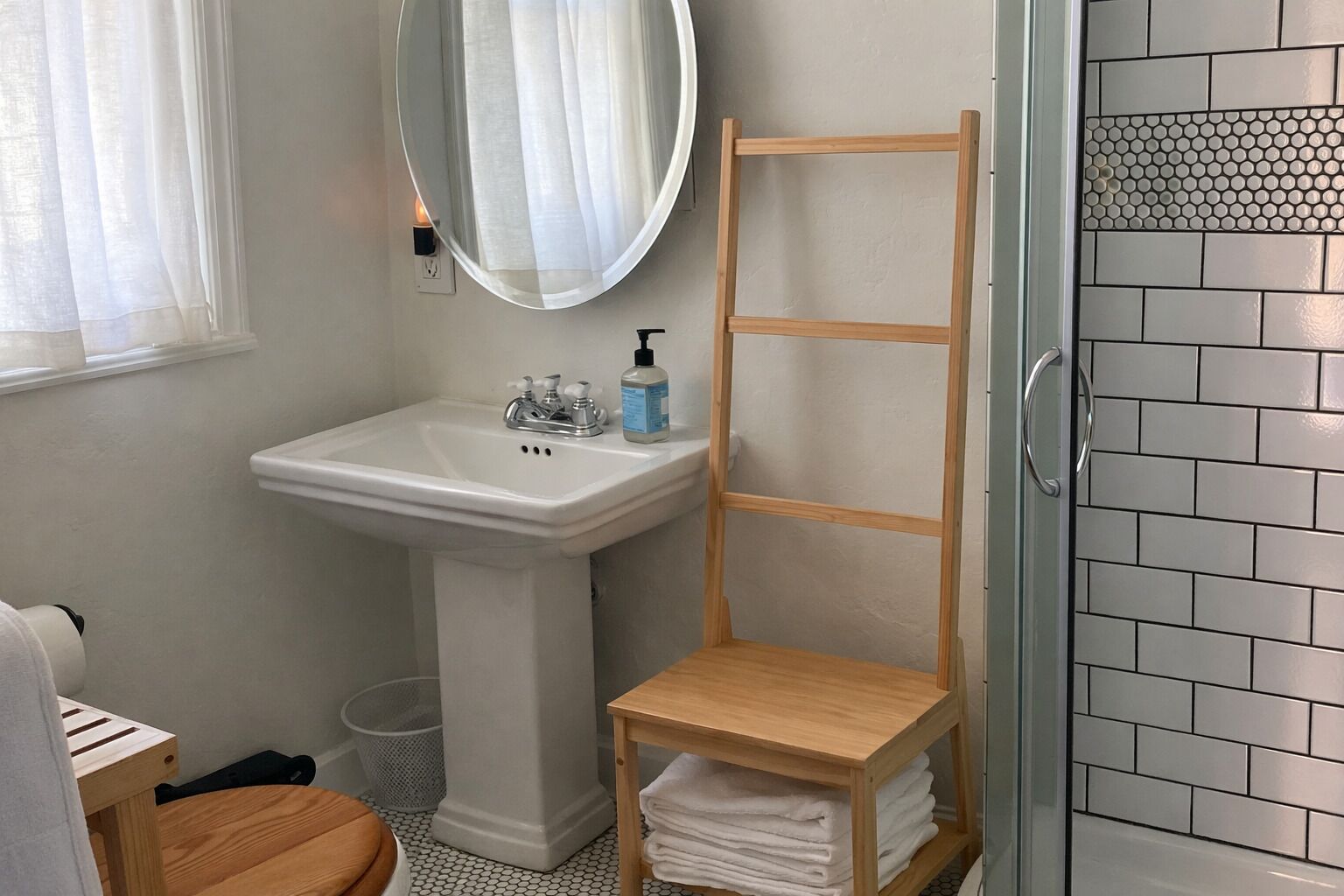 Un baño con lavamanos pedestal, estante de madera, espejo y zona de ducha con azulejos.