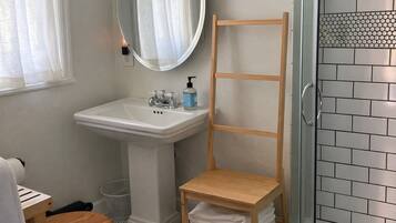 Un baño con lavamanos pedestal, estante de madera, espejo y zona de ducha con azulejos.
