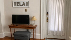 A room with a television on a wooden table, a small lamp, a grey ottoman, a framed word 'RELAX' on the wall, and a patterned rug on the floor.