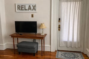 A room with a television on a wooden table, a small lamp, a grey ottoman, a framed word 'RELAX' on the wall, and a patterned rug on the floor.