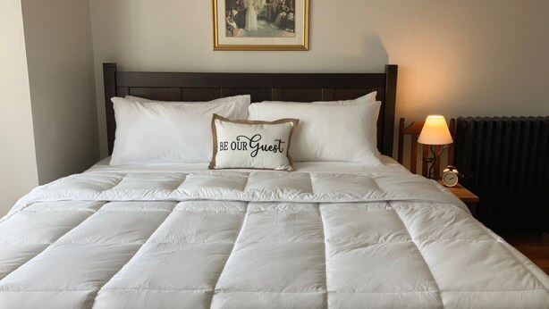 A neatly made bed with a quilted duvet, a pillow with 'BE OUR Guest' text, a framed picture on the wall, and a bedside table with a lamp and an alarm clock.