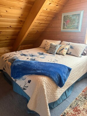 A cozy attic bedroom with a wooden ceiling, a bed with floral bedding, and a painting on the wall.