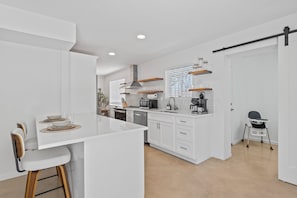 A modern kitchen with white cabinetry, a central island, and a dining area with bar stools. - Summer Retreat with Pool & Views - Sleeps 8! (Tucson)