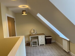 Ein kleines Schlafzimmer im Dachgeschoss mit sanfter Beleuchtung, einer geneigten Decke, einem weißen Schminktisch mit Spiegel, einer Holztür und einem Heizkörper.