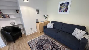 En stue med en mørkeblå sofa, et mønstret tæppe, en bogreol, en lille sort stol, et træskrivebord og et billede i ramme på væggen.