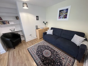 A living room with a dark blue sofa, a patterned rug, a bookshelf, a small black chair, a wooden desk, and a framed picture on the wall. - Ancluaran (The Thistle) newly renovated one bedroom house 2+2 (Fort Augustus)