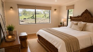 A bedroom with a large bed, a bedside table, a nightstand, a window with a view of the outdoors, and a wall-mounted lamp.