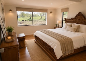 A bedroom with a large bed, a bedside table, a nightstand, a window with a view of the outdoors, and a wall-mounted lamp.