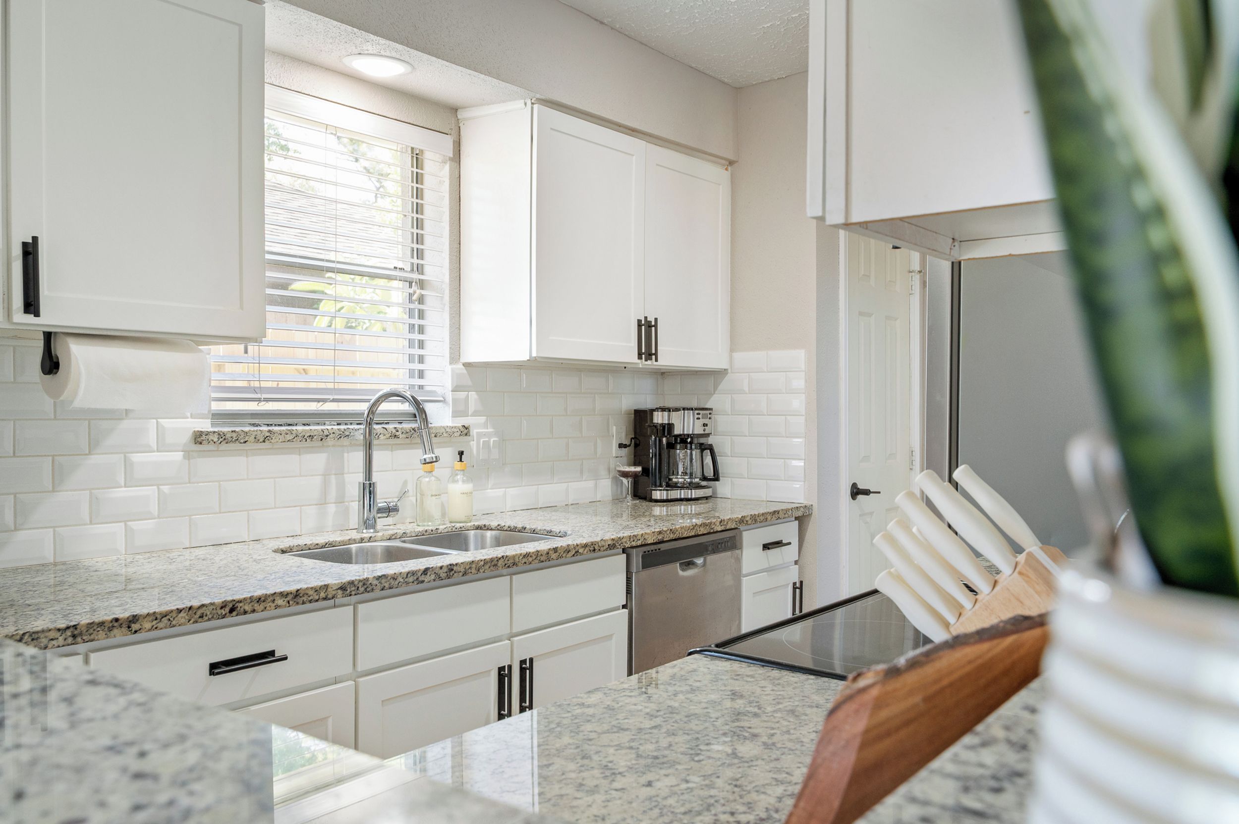 A modern kitchen with white cabinetry, granite countertops, a built-in dishwasher, and a coffee maker.