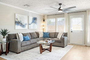 Ein Wohnzimmer mit einem grauen Ecksofa, zwei abstrakten Gemälden, einem Glastisch und einem Deckenventilator.
