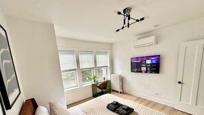 A bedroom with a bed, a TV mounted on the wall, a radiator, and a window with blinds.