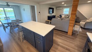 Una cocina moderna con una isla central, una zona de comedor con mesa y sillas, una sala de estar con sofá y televisor, y una escalera que sube a un nivel superior.