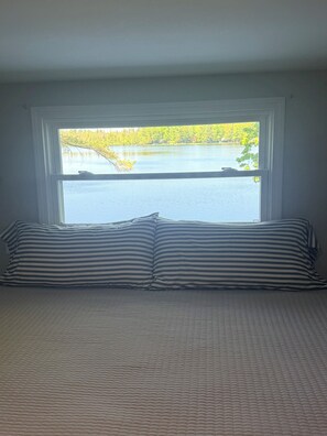 A bedroom with a large window, a bed with striped bedding, and a view of a lake and trees. - Lakefront Cottage Near Torch Lake • Private Dock •Kayaks •Sunsets (Kalkaska)