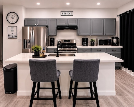 A modern kitchen with grey cabinets, stainless steel appliances, a large island with two bar stools, and a clock on the wall.. Cozy 2BR Houston Home Steps to MetroRail & Downtown Access & WORLD CUP ACCESS
