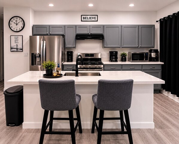 Una cocina moderna con armarios grises, electrodomésticos de acero inoxidable, una isla grande con dos taburetes de bar y un reloj en la pared.