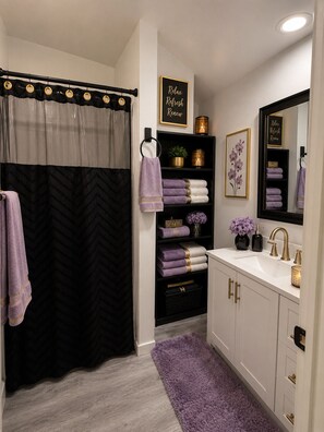 A bathroom with a black and white chevron curtain, a purple towel, a shelf with folded towels, a mirror, and a sink with gold fixtures. - PRIVATE GATED 2BR Home Steps 2 MetroRail & Minutes to Downtown, MedCenter, &FIFA (Houston)