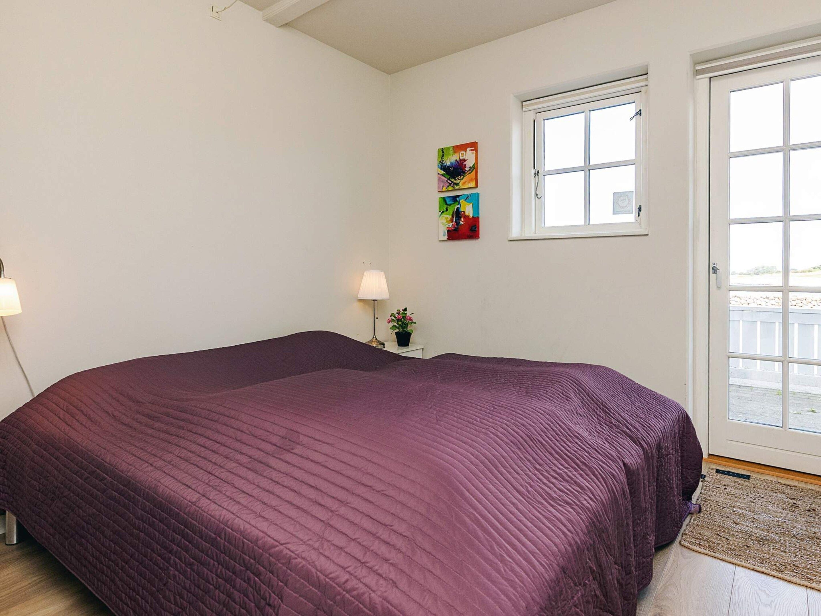 Ein Schlafzimmer mit einem großen Bett, einem Fenster, einer Tür und einem kleinen Tisch mit Lampe und Pflanze.