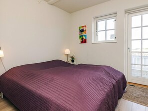 Une chambre à coucher avec un grand lit, une fenêtre, une porte et une petite table sur laquelle se trouvent une lampe et une plante.
