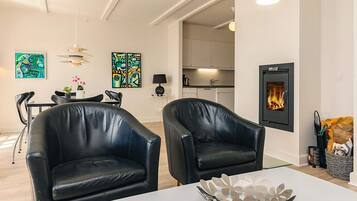Uma sala de estar moderna com duas poltronas de couro preto, uma mesa branca com uma tigela decorativa de metal, uma lareira e uma área de cozinha ao fundo.