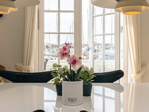 Une table sur laquelle est posé un vase blanc contenant des fleurs roses et des feuilles vertes, placée devant une fenêtre donnant sur des bateaux et des bâtiments.