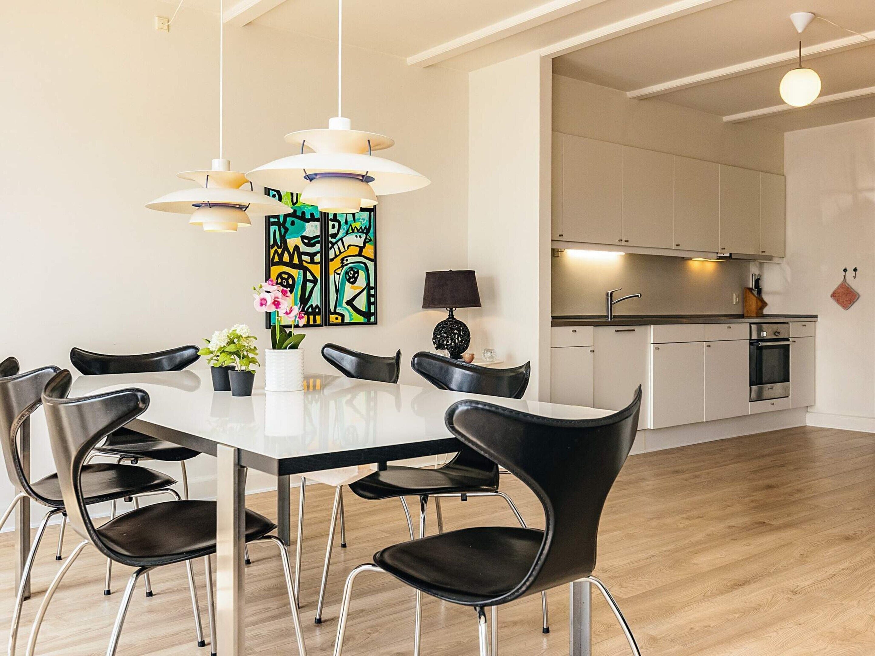 Uma sala de jantar moderna com uma mesa branca, cadeiras pretas e iluminação pendente.