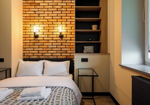 Ein Hotelzimmer mit Backsteinwand, einem Bett mit weißer Bettwäsche, einem kleinen Tisch und einem Regal mit Körben.