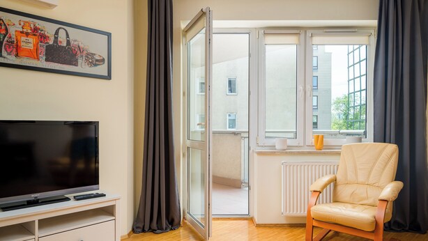 Een moderne woonkamer met een flatscreen televisie, een beige fauteuil, een ingelijste foto aan de muur en een groot raam met gordijnen.
