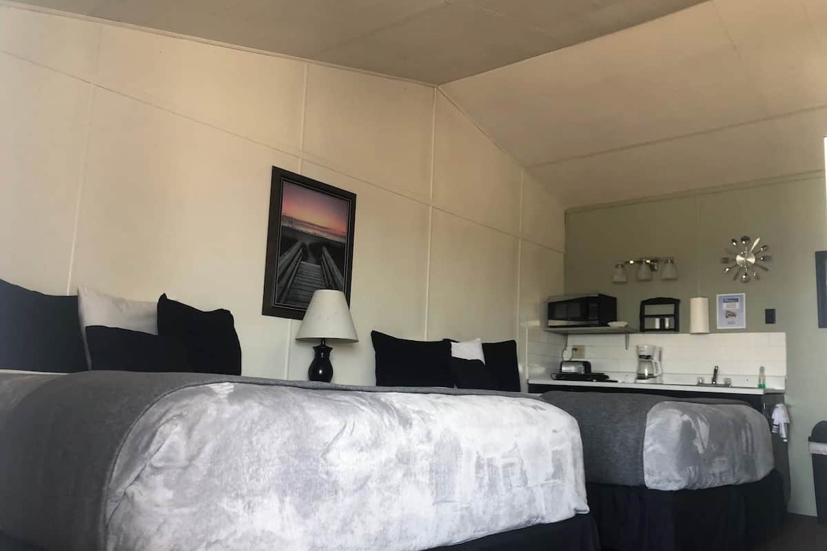 A hotel room with two beds, a desk, a microwave, a coffee maker, and a picture of a sunset over a pier.