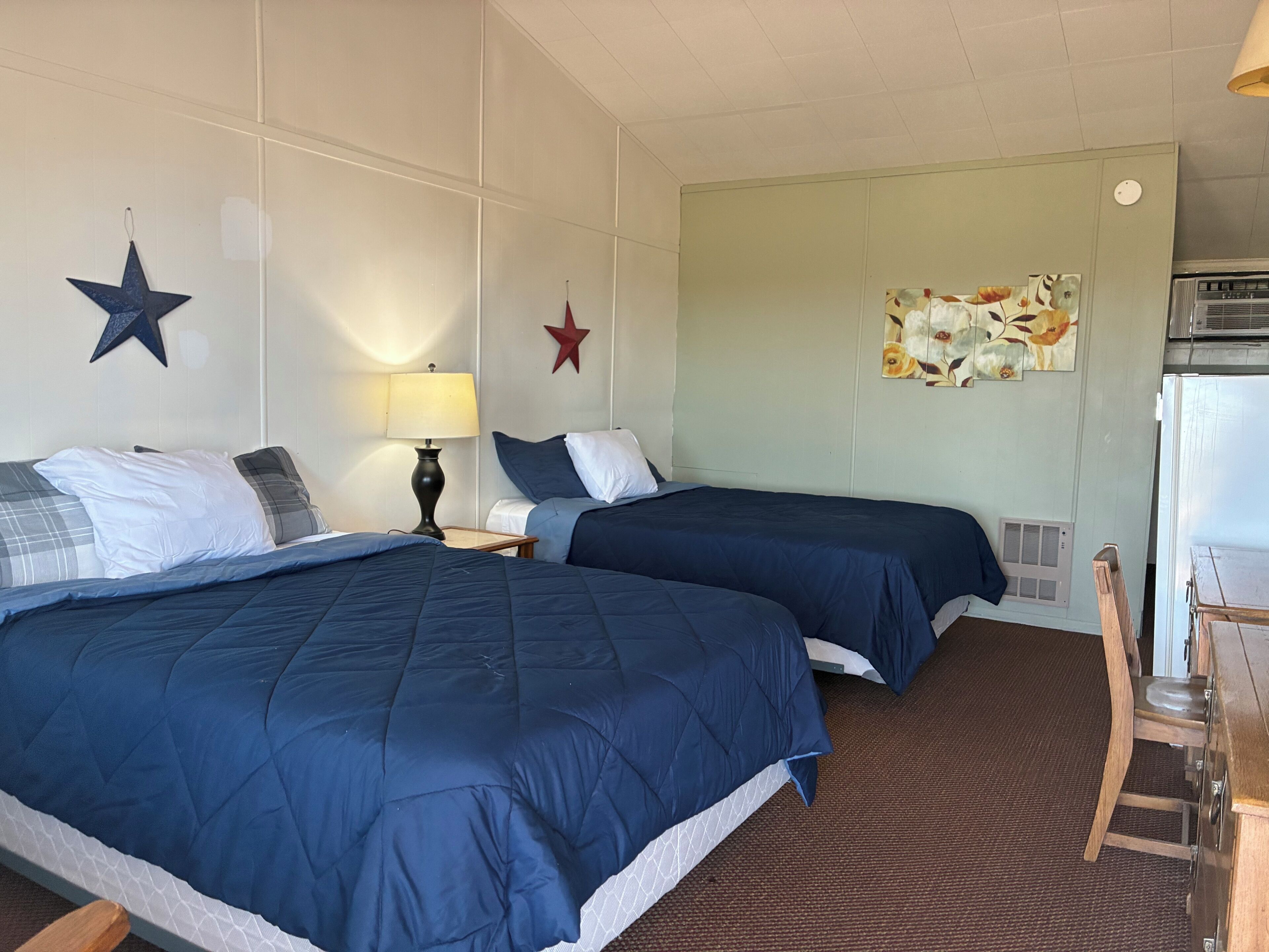 A hotel room with two beds, a desk, a lamp, and a refrigerator.