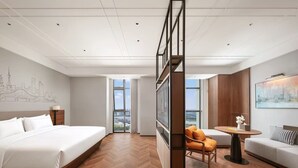 Habitación de hotel con una cama grande, un sofá, una mesita y vistas a la ciudad a través de amplios ventanales.