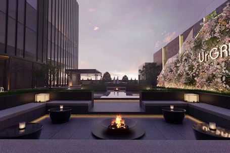 A modern outdoor seating area with a central fire feature, surrounded by minimalist tables and benches, set against a backdrop of contemporary architecture and a floral wall.. UrCove Foshan Tanzhou Exhibition Center