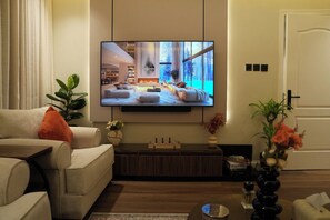 A modern living room with a flat-screen TV mounted on the wall, a sofa, a coffee table, and a decorative vase with flowers. - Cozy 1-bedroom apartment in Almlaqa Riyadh with coffee bar (Riyadh)