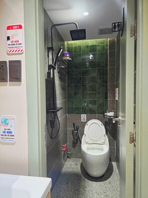 Un bagno piccolo con il water, una doccia e un lavandino. Sui muri ci sono delle indicazioni, tra cui un cartello con scritto 'Chiudere l'acqua quando non serve'.